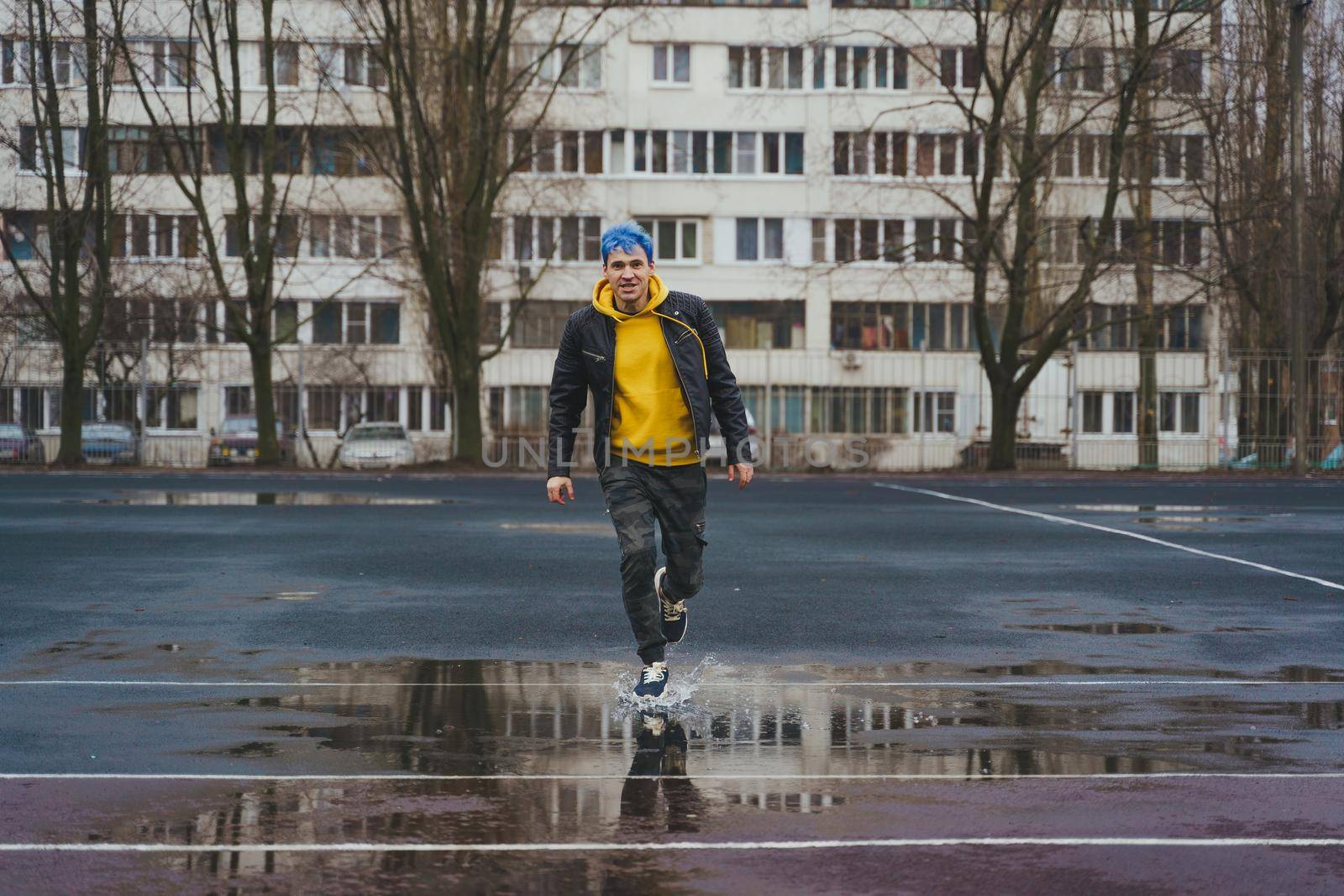 Young man runs through puddle on sports stadium on background of high-rise building. Happy guy with blue hair having fun on city street in springtime