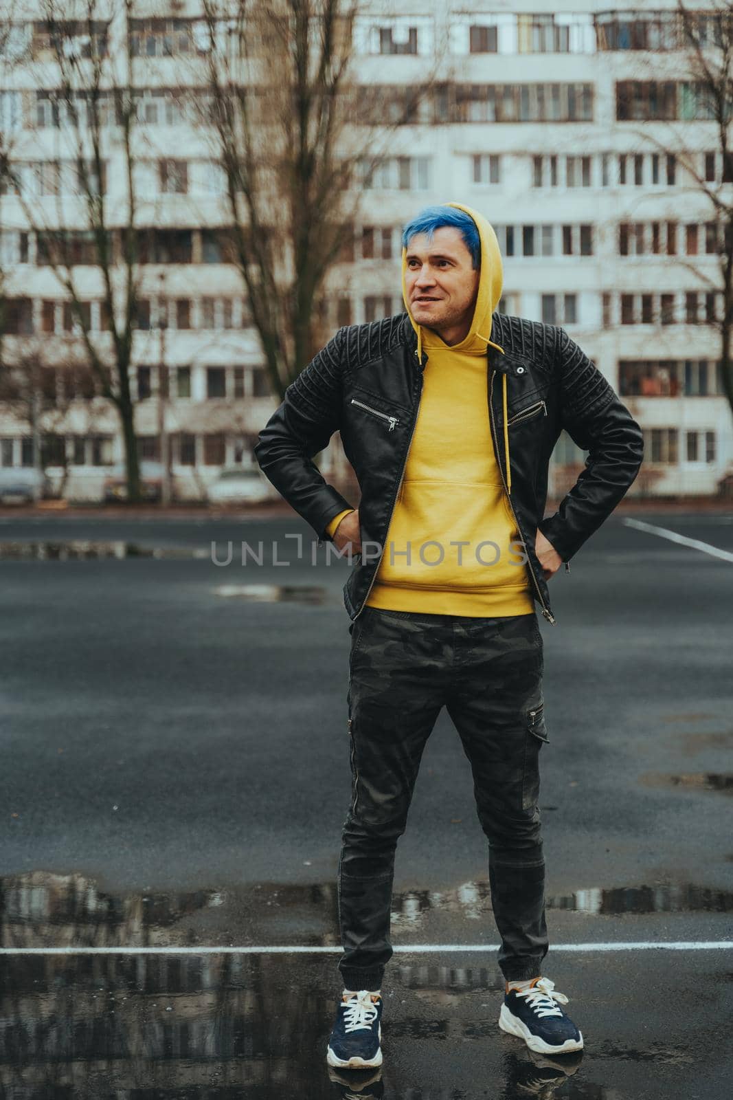 Young man standing on sports stadium on background of high-rise building. Handsome guy with blue hair posing on city street in springtime. by epidemiks