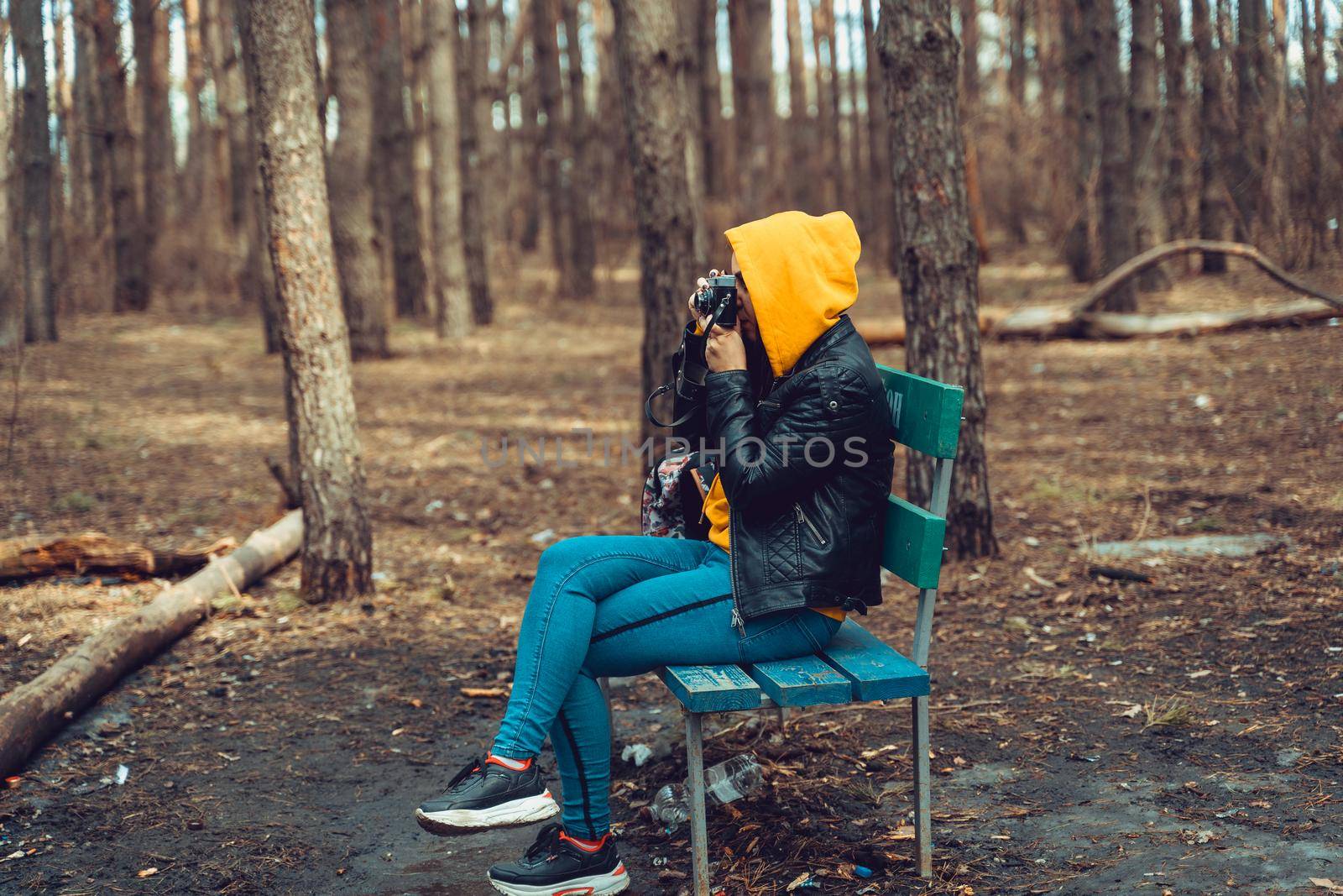 Young woman in casual clothes sitting on bench and photographing on old photo camera in forest. Female resting and taking photos with old camera in early spring. by epidemiks