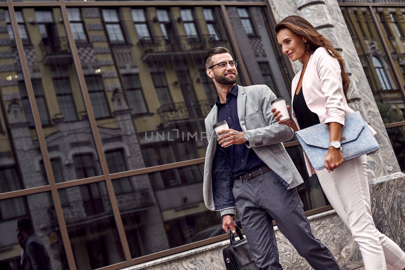 Hurry for meeting. Two young business people walking outside on the city street drinking hot coffee talking smiling joyful close-up by friendsstock