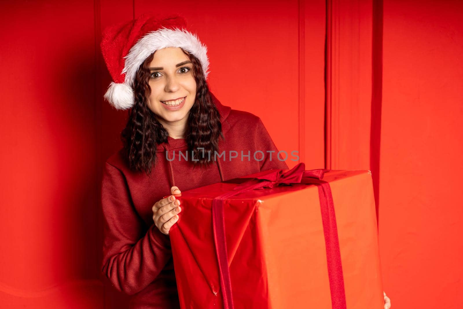 Young woman in Santa Claus hat holds big gift, standing near red wall. Happy female in Christmas hat with box of gift. Concept of holidays, presents and good mood