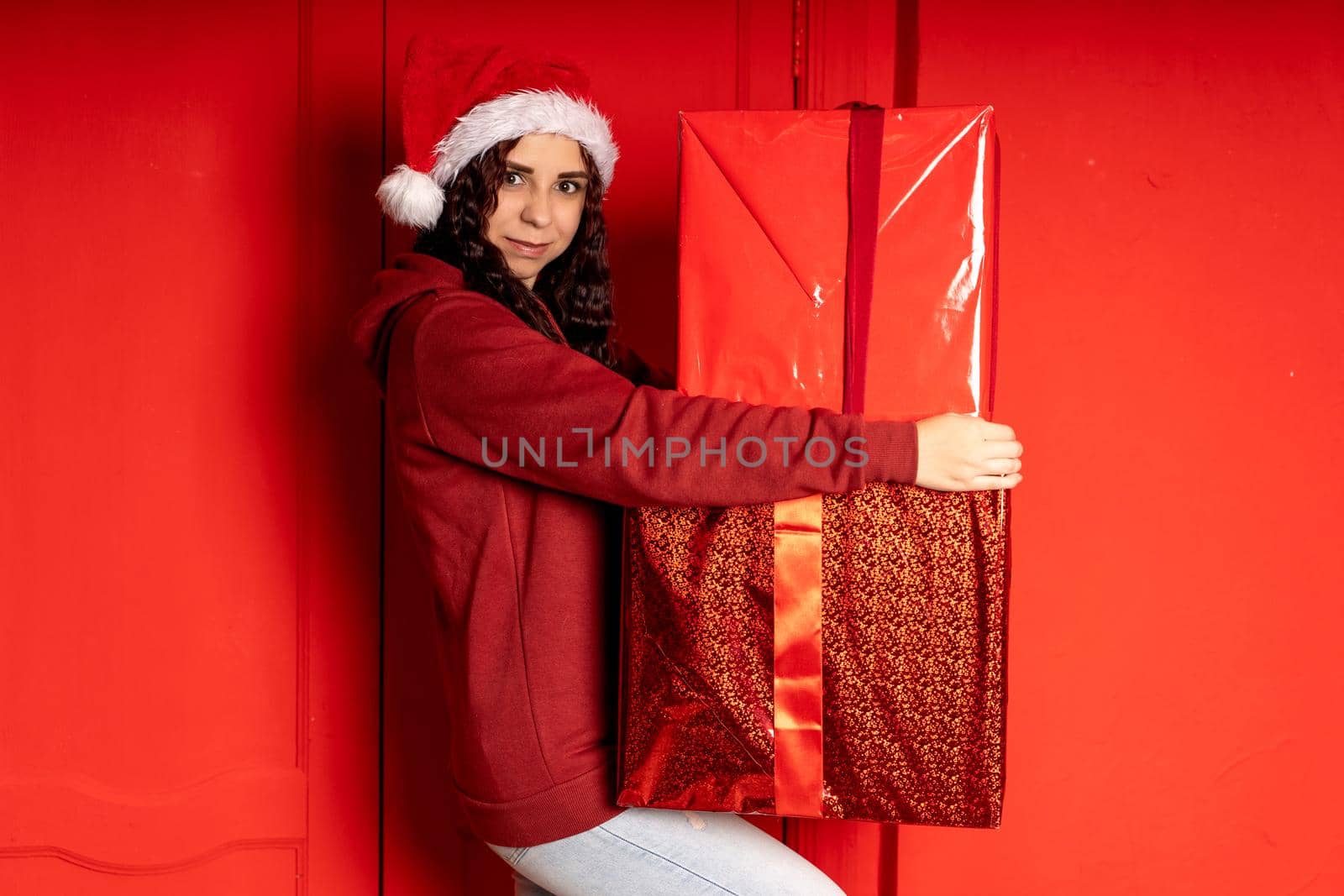 Young woman in Santa Claus hat holds big gifts, standing near red wall. Happy female in Christmas hat with boxes of gift. Concept of holidays, presents and good mood