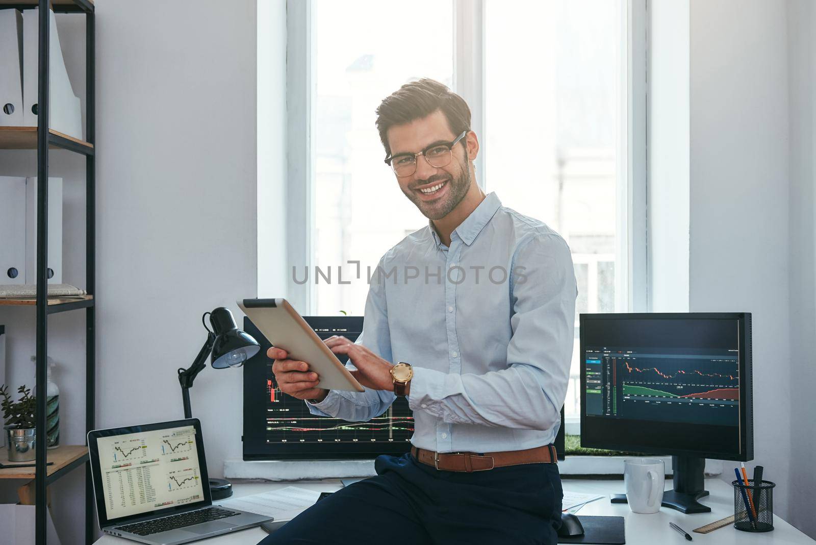Business success. Cheerful young trader in formal wear is holding digital tablet and looking at camera with smile while standing in front of computer screens with trading charts in the office. by friendsstock
