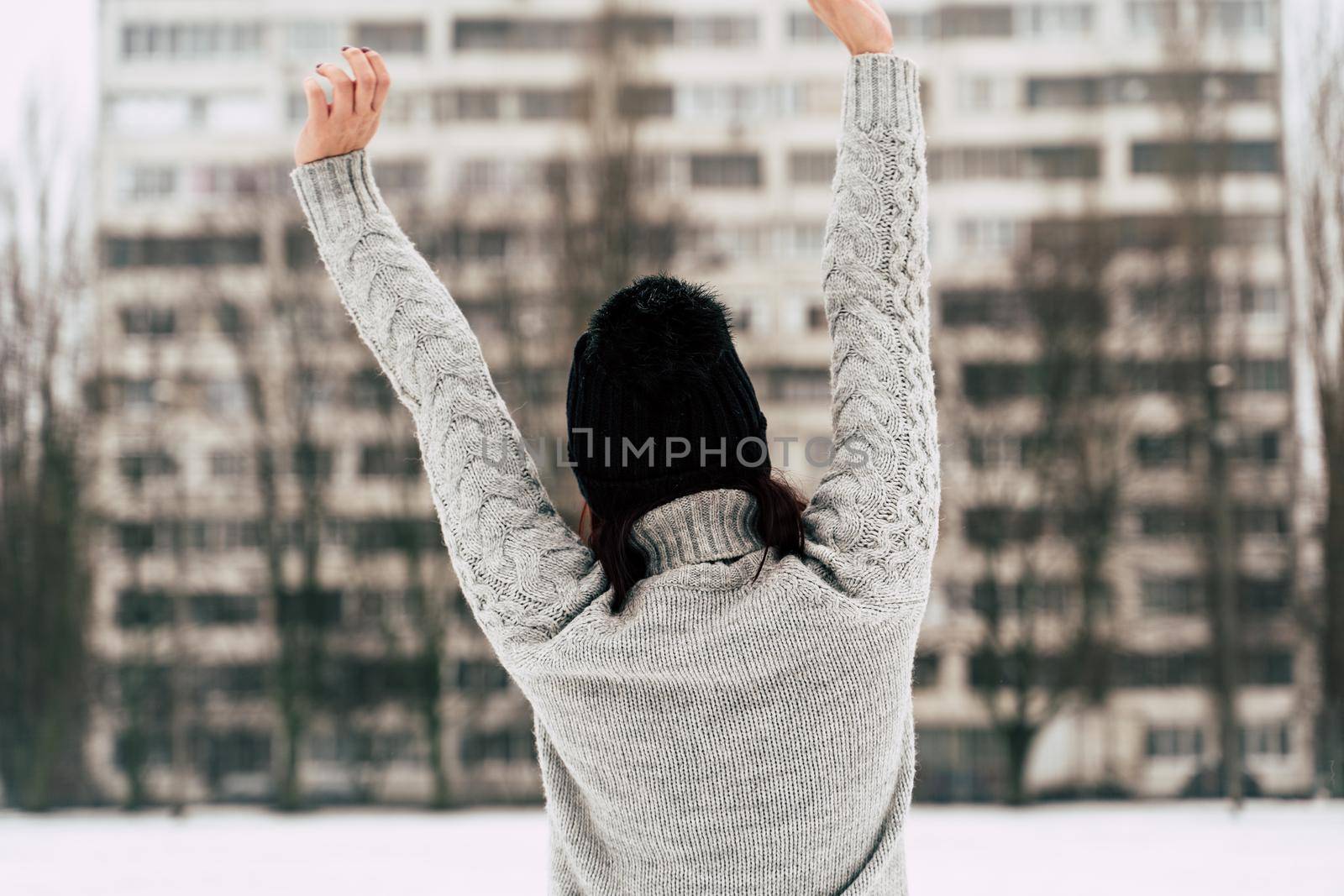 Rear view of young woman in gray knitted sweater and hat standing on street in winter season. Female looking at high-rise building and stretching her hands in cloudy weather. by epidemiks
