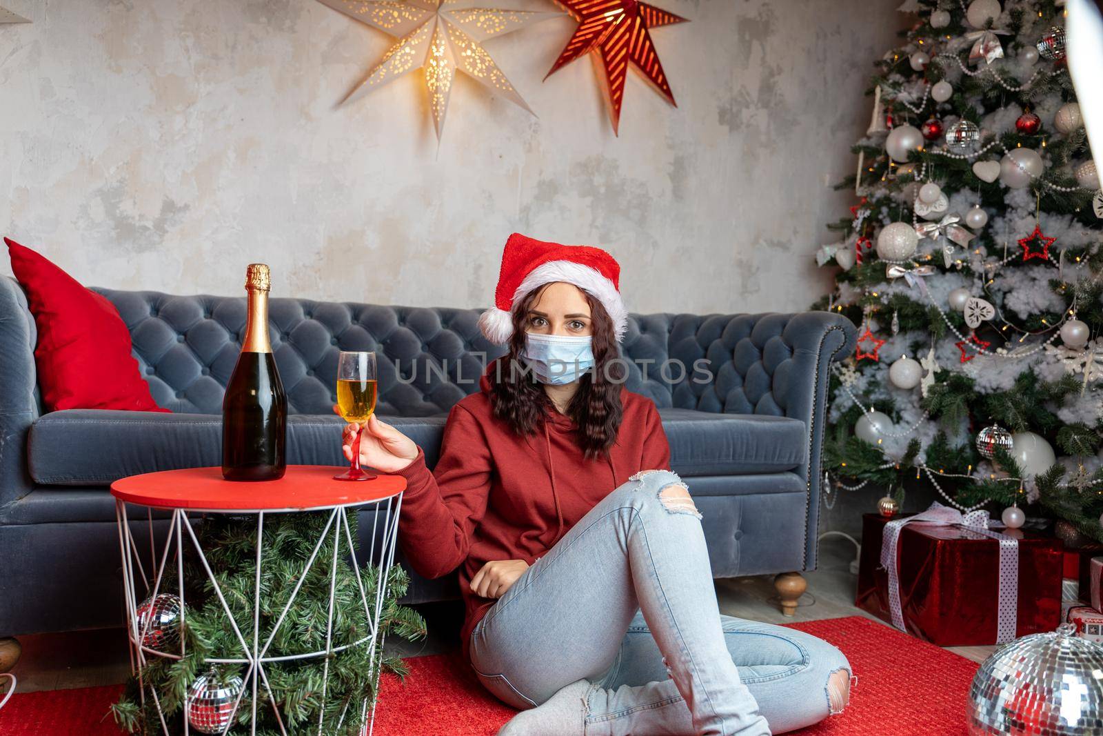 Young woman in medical mask and Santa Claus hat sitting on floor near sofa in room. Alone charming brunette in protective mask celebrating of safe Christmas during coronavirus pandemic. by epidemiks