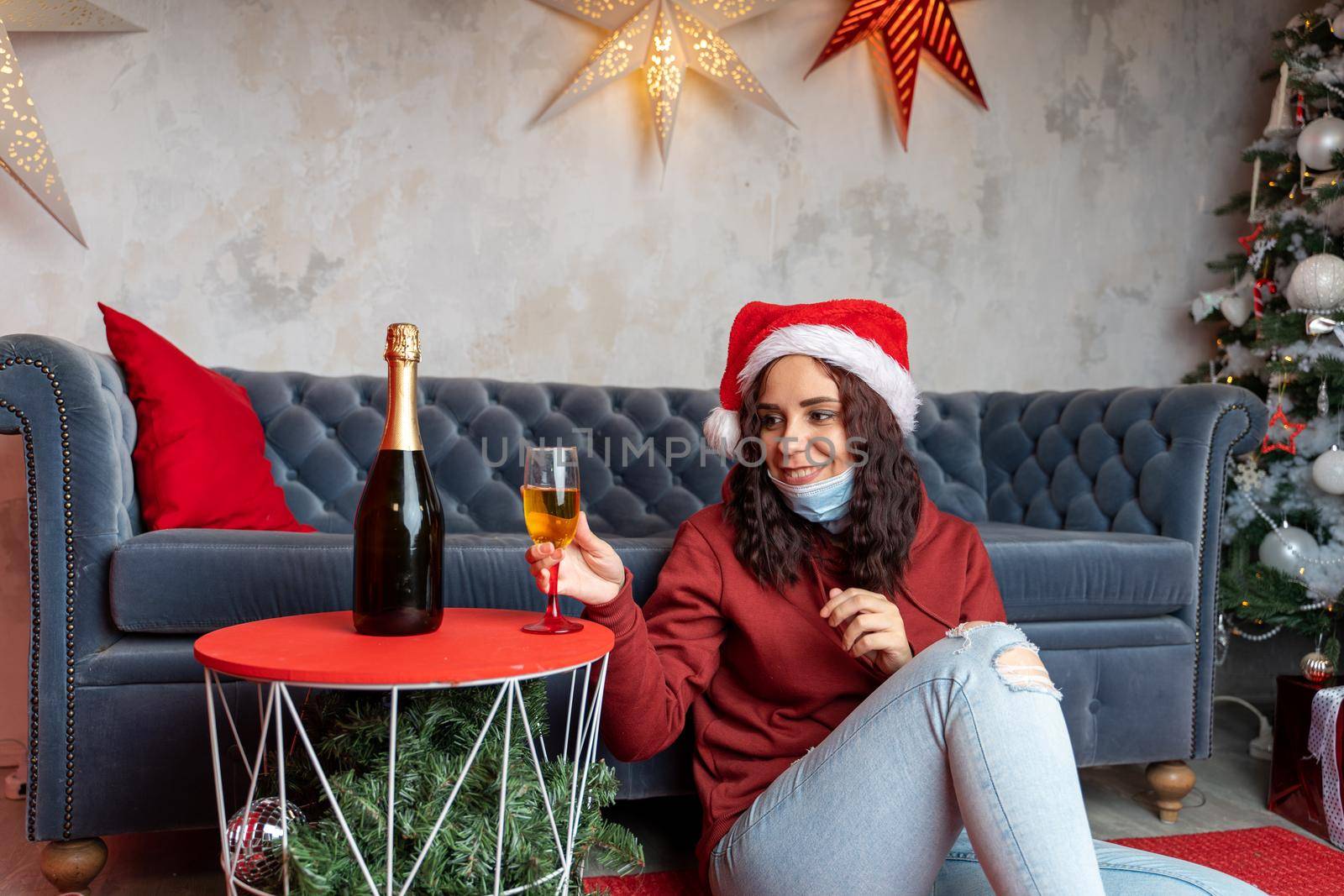 Young woman in medical mask on beard and santa hat takes glass of champagne, sitting on floor near sofa in room. Charming brunette smiles, celebrating of safe Christmas during coronavirus pandemic