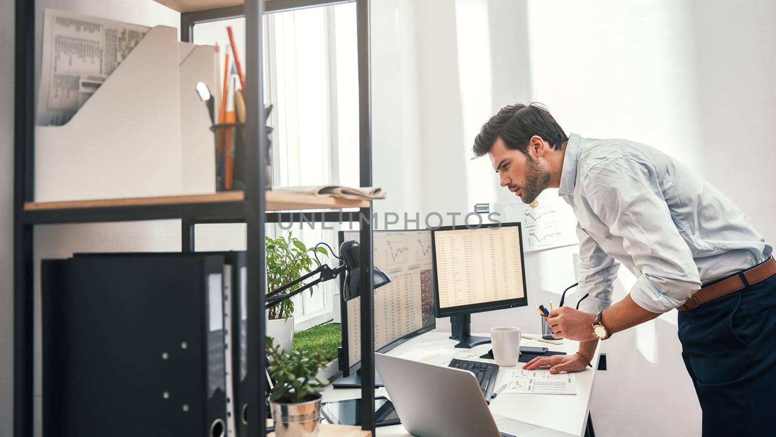 Busy day. Young successful trader or businessman in formal wear looking at computer screens with charts while standing at his modern office. Stock broker. Forex market. Trade concept