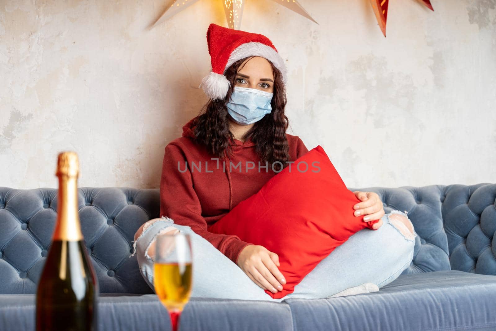 Young woman in medical mask and Santa Claus hat sitting on sofa in room. Alone charming brunette in protective mask celebrating of safe Christmas during coronavirus pandemic