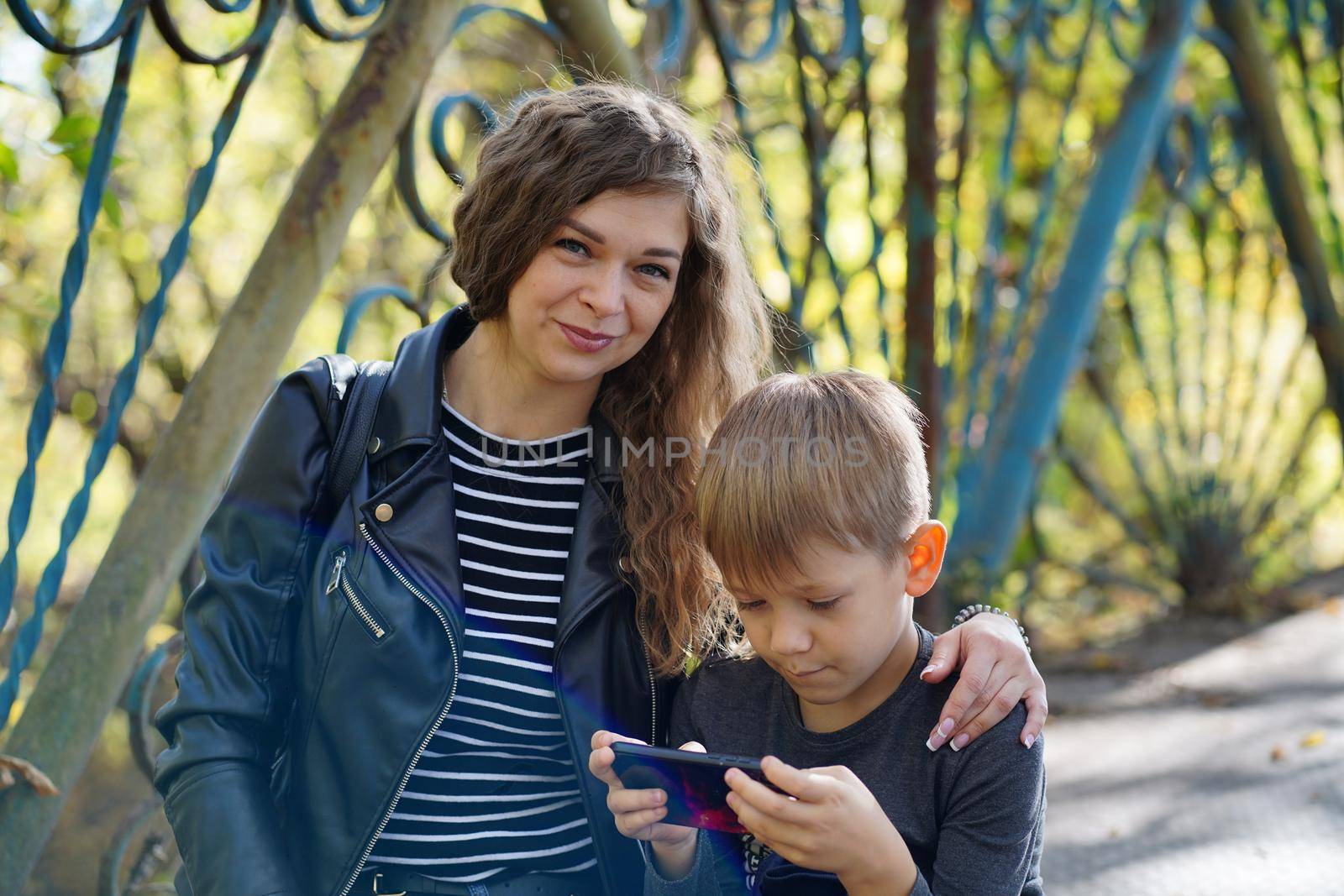 A focused boy is playing a game on a smartphone in the park. A serious focused school age boy in casual clothes is sitting on a bench and together with his mother and playing a game on a mobile phone by epidemiks