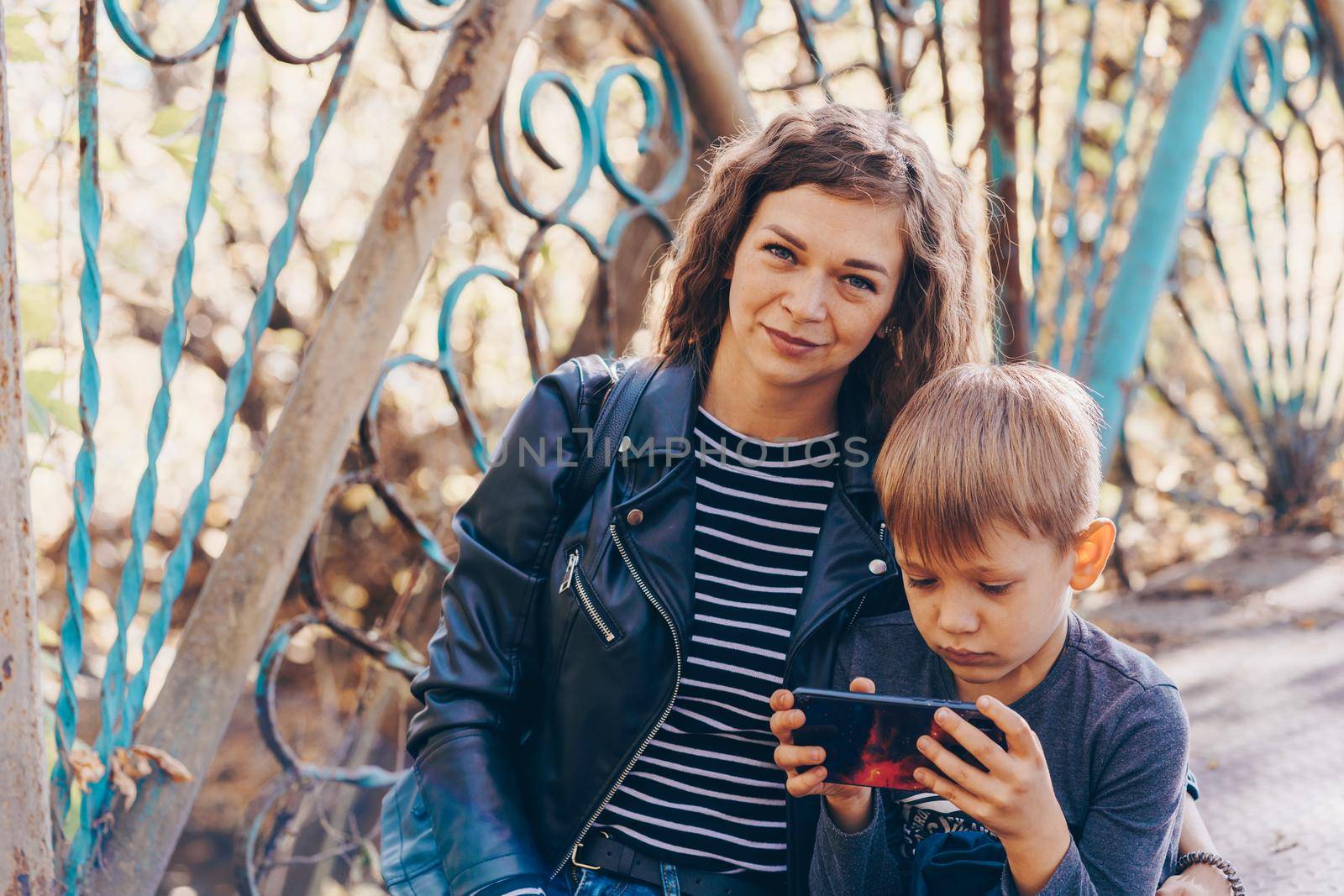 A focused boy is playing a game on a smartphone in the park. A serious focused school age boy in casual clothes is sitting on a bench and together with his mother and playing a game on a mobile phone by epidemiks