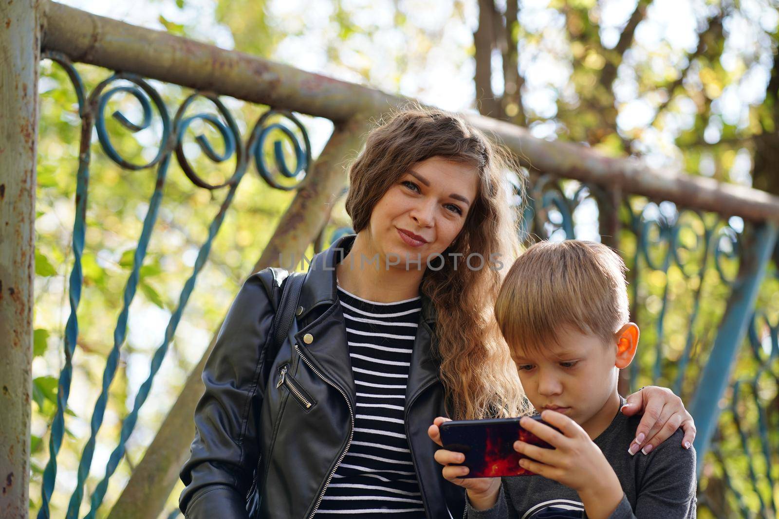 A focused boy is playing a game on a smartphone in the park. A serious focused school age boy in casual clothes is sitting on a bench and together with his mother and playing a game on a mobile phone.
