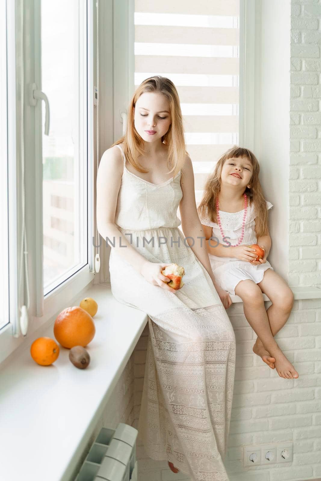 Two girls in white dresses are eating fruit while sitting by the window in a white kitchen. by Lena_Ogurtsova