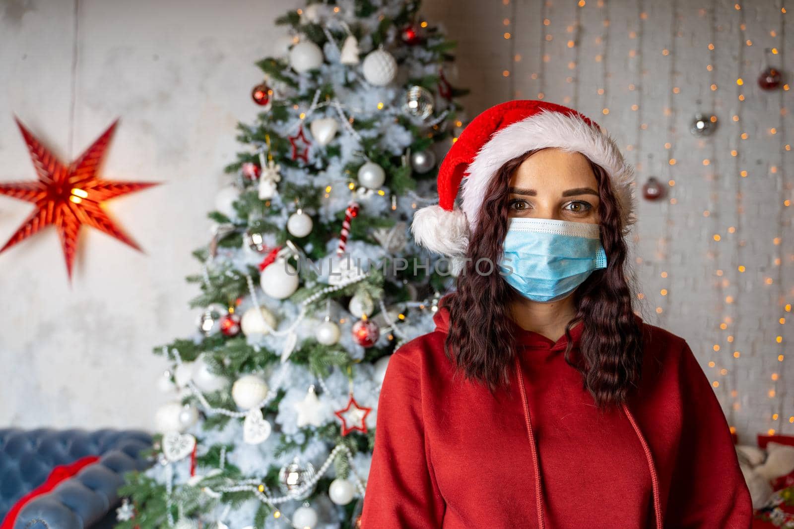 Young woman in medical mask and santa hat on background of Christmas tree. Charming brunette in protective mask celebrating of safe Christmas during coronavirus pandemic. by epidemiks