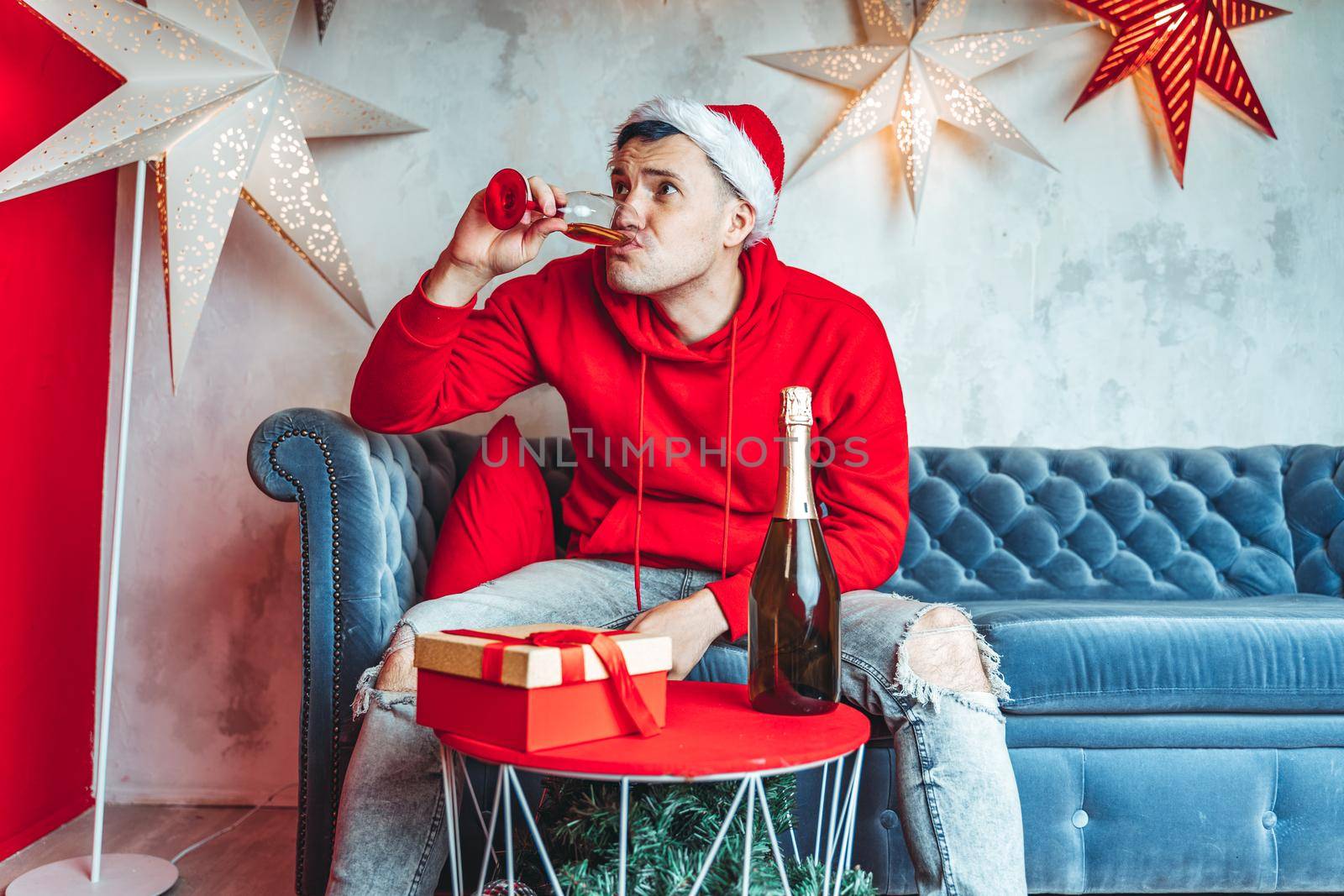 Young man in Santa hat celebrating Christmas in solitude at home. Adult guy rests, sitting on sofa in room. Concept of holidays and good mood