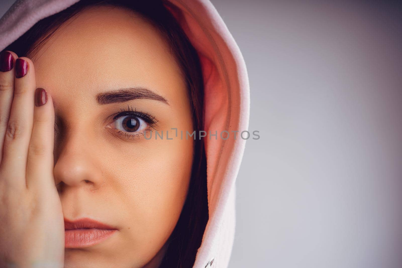 Portrait of young woman closing her eye with hand. Close up of pretty female in hood hiding part of face behind hand