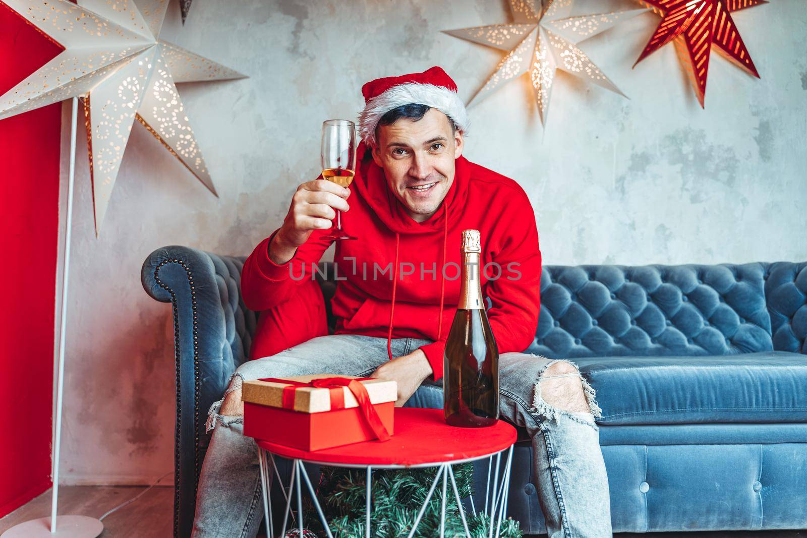 Young man in Santa hat celebrating Christmas in solitude at home. Adult guy rests, sitting on sofa in room. Concept of holidays and good mood. by epidemiks