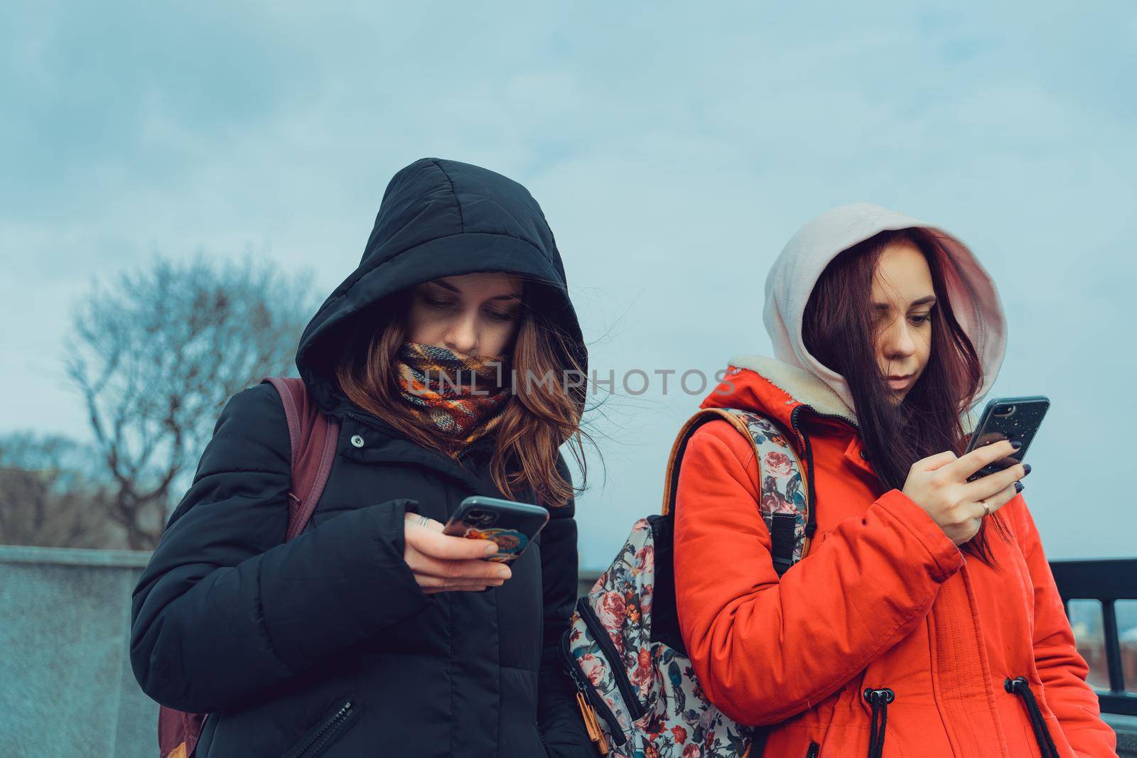 Close up of young women browsing smartphone in city park. Pretty females in hoods using mobile phone. by epidemiks