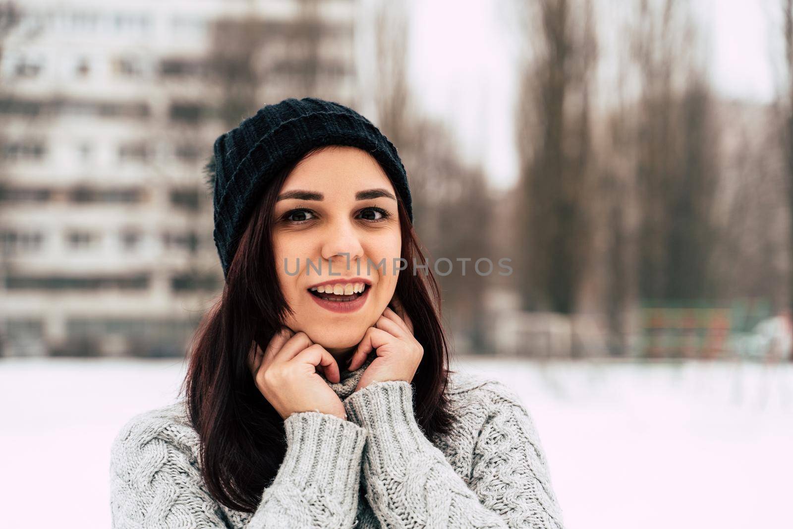 Portrait of young woman in gray knitted sweater and hat on background of high-rise building. Close up of pretty female posing and looking at camera on walk in winter season