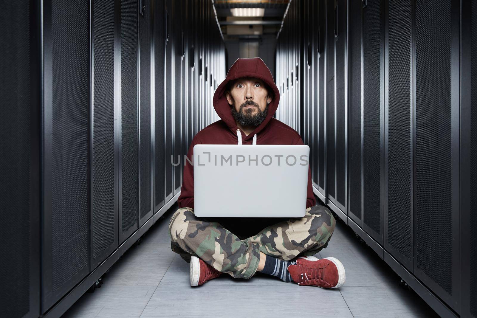 The hacker in a hood sits with the computer among servers, the burglar. Storage of data, cloudy services, system breaking, attack of hackers by vladimirdrozdin