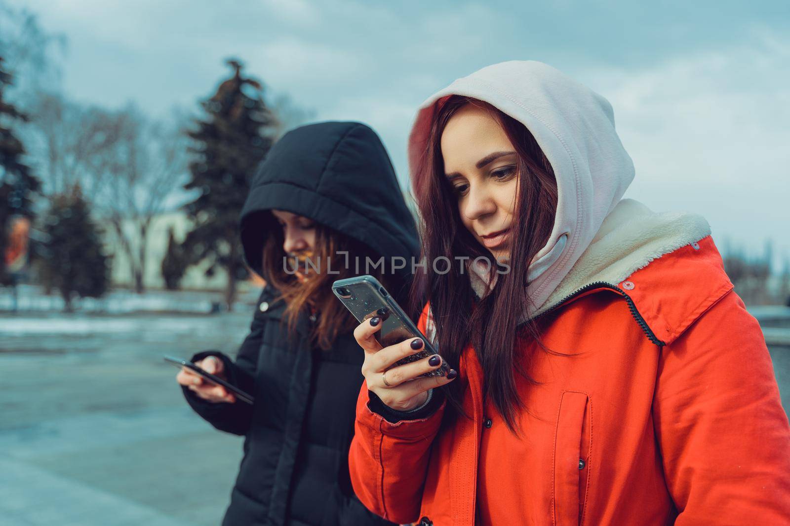 Close up of young women browsing smartphone in city park. Pretty females in hoods using mobile phone
