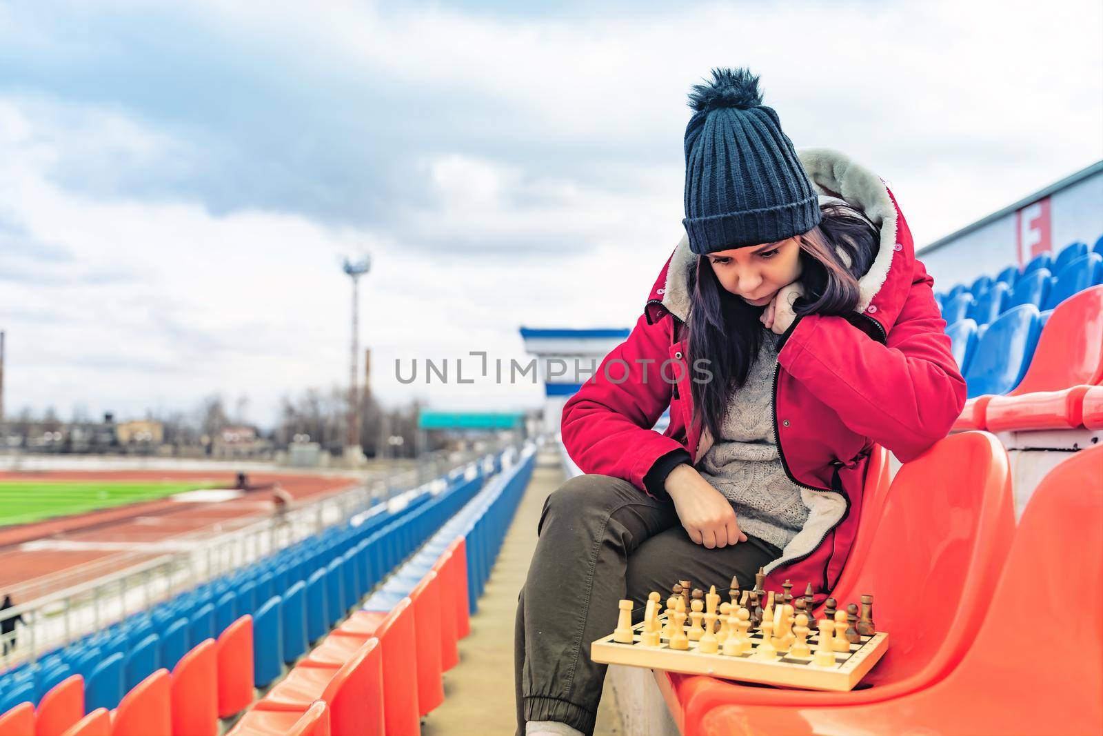 Young woman in winter clothes plays chess, sitting on stadium bleachers alone. Female in black cap with chess on sports stadium in cloudy weather. by epidemiks