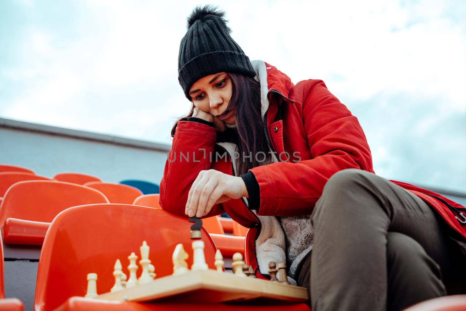 Young woman in winter clothes plays chess, sitting on stadium bleachers alone. Female in black cap with chess on sports stadium in cloudy weather