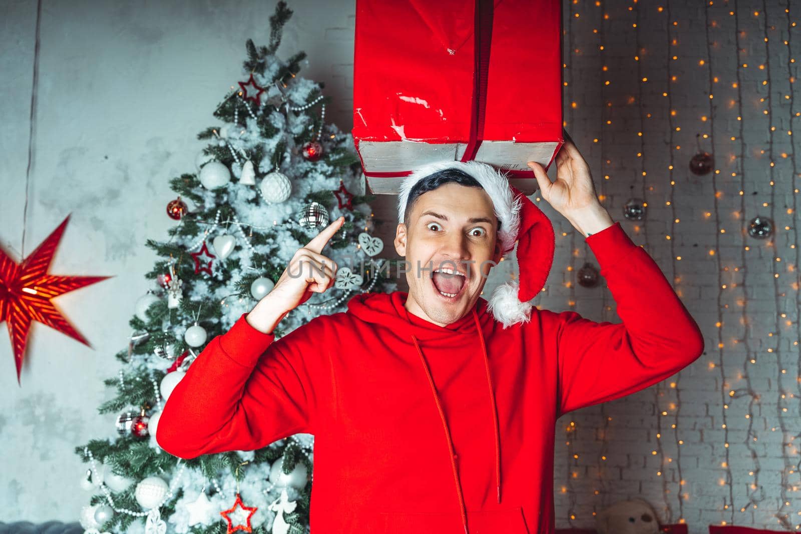 Young handsome man in Santa Claus hat dancing with big gift on background of Christmas tree. Happy guy dances, rejoicing present. Concept of holidays, presents and good mood