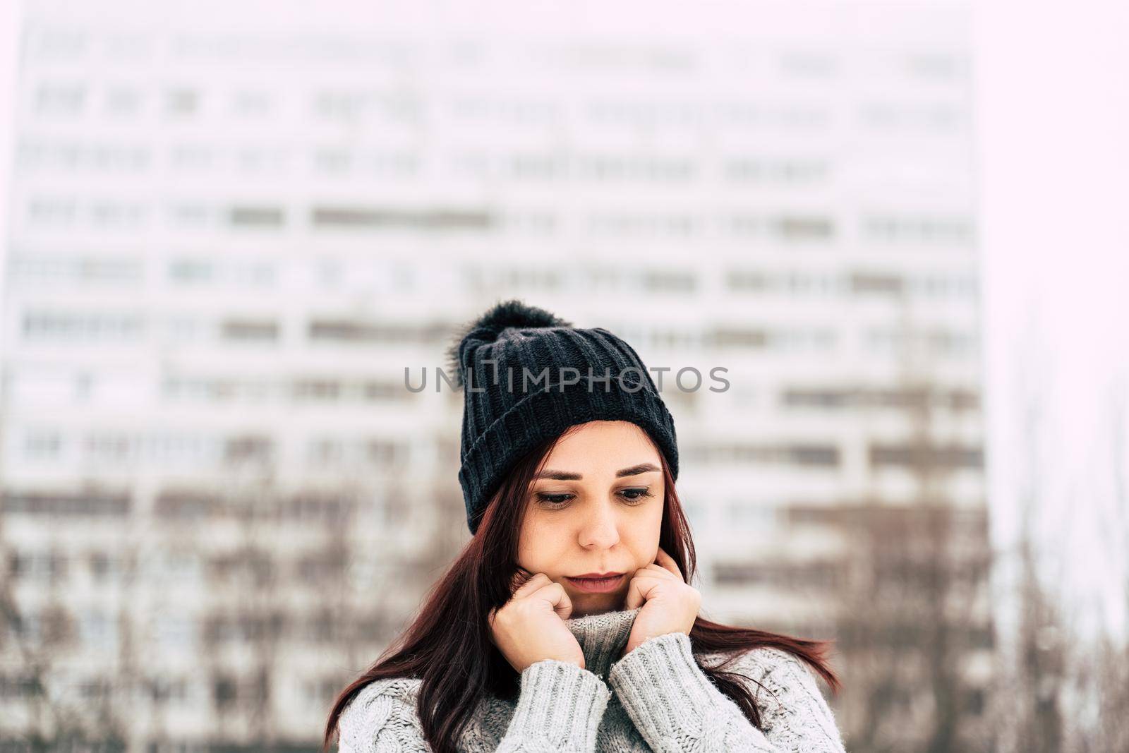 Portrait of young woman in gray knitted sweater and hat on background of high-rise building. Close up of pretty female posing and looking down on walk in winter season