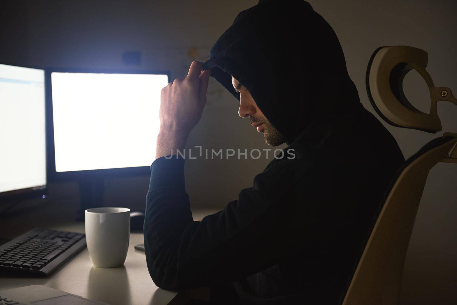 Computer hacker. Young man in black hoodie using multiple computers for stealing data while sitting in dark room. Binary code. Cyber attack. Cyber security