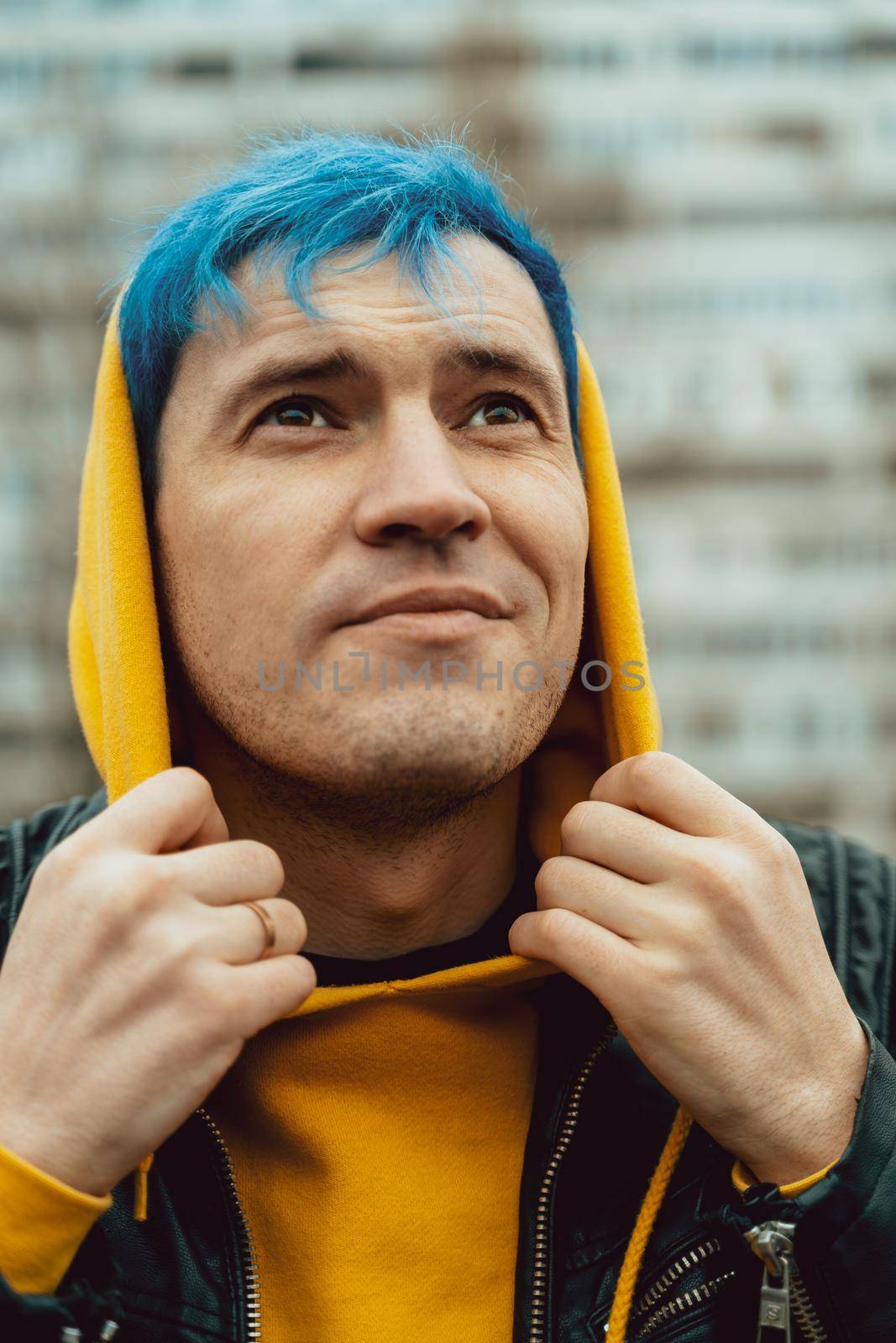 Portrait of young man in hood on background of high-rise building. Handsome guy with blue hair posing on city street in springtime. by epidemiks
