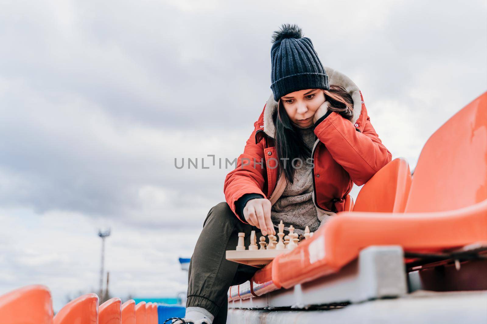 Young woman in winter clothes plays chess, sitting on stadium bleachers alone. Female in black cap with chess on sports stadium in cloudy weather. by epidemiks
