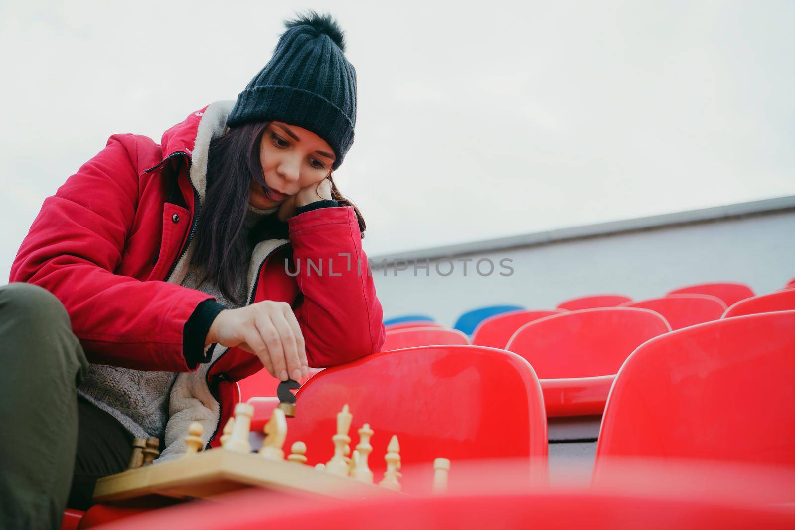 Young woman in winter clothes plays chess, sitting on stadium bleachers alone. Female in black cap with chess on sports stadium in cloudy weather. by epidemiks