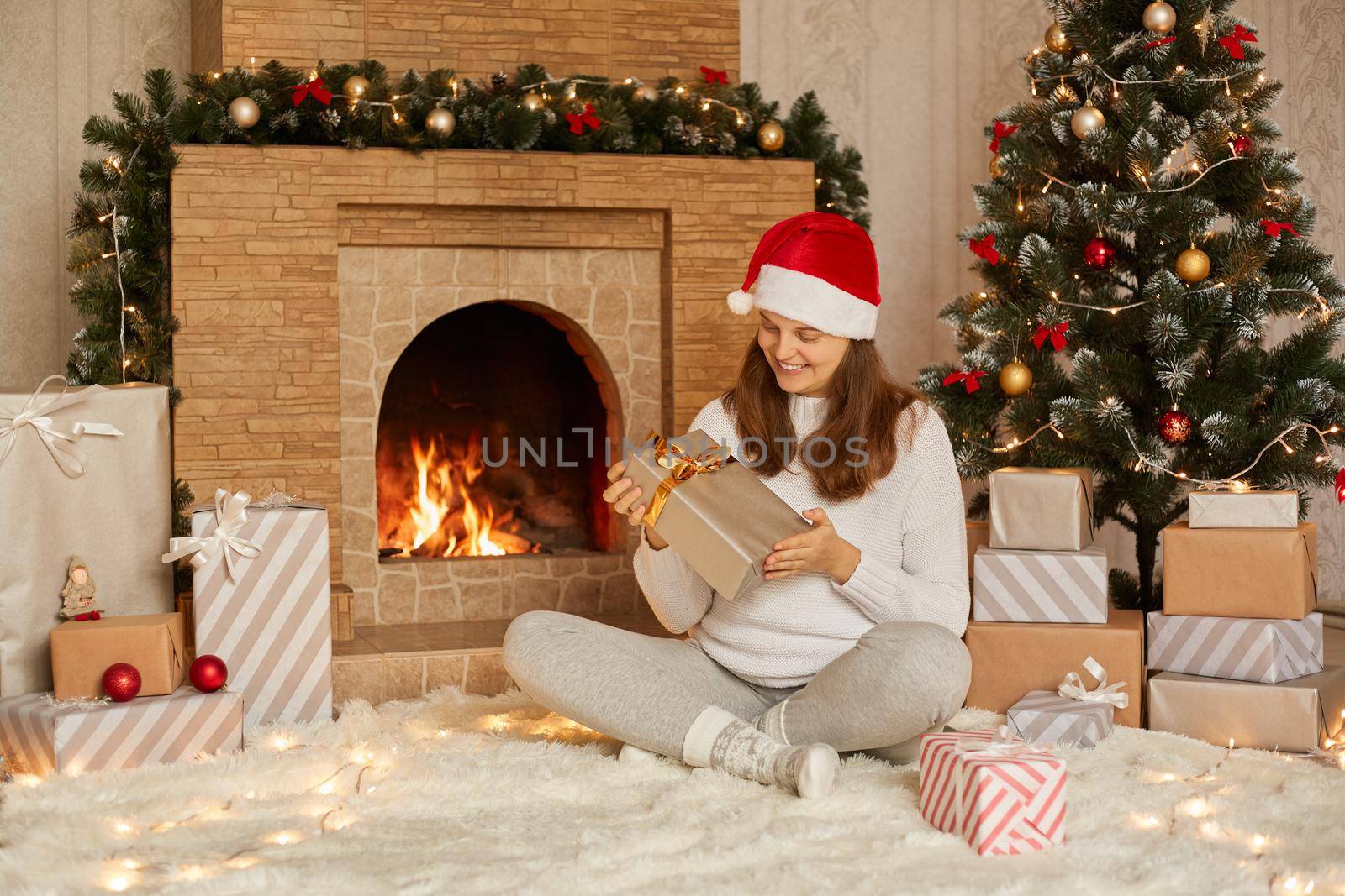 Lovely woman opening gift on Christmas, sitting on floor near fireplace and tree, smiling female wearing santa hat, white sweater and warm socks, sitting with crossed lags and holding present box.