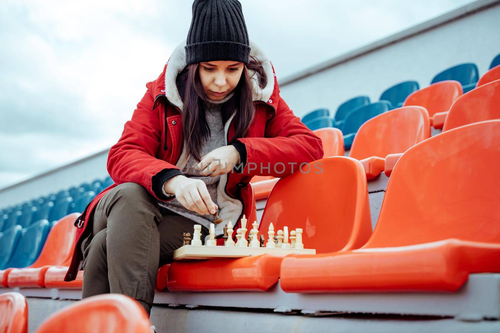 Young woman in winter clothes plays chess, sitting on stadium bleachers alone. Female in black cap with chess on sports stadium in cloudy weather