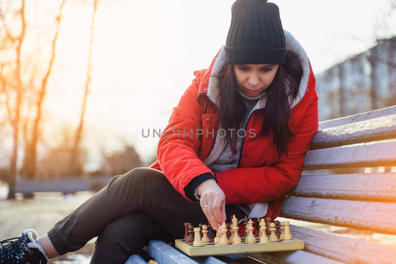 Young woman in winter clothes plays chess, sitting on bench in city park. Female in black cap with chess on wooden pew in cloudy weather