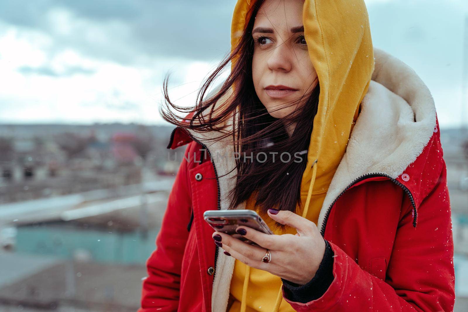 Portrait of young woman with mobile phone on roof of building. Brunette in yellow hoodie and red jacket browsing smartphone in cloudy weather
