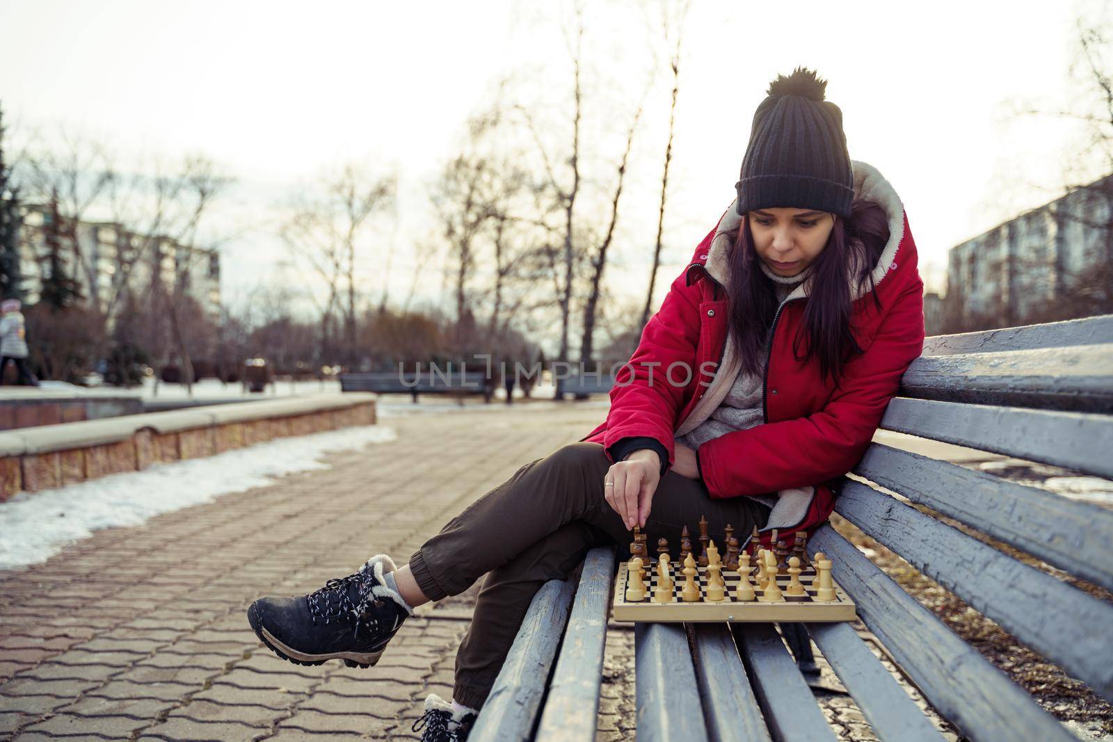 Young woman in winter clothes plays chess, sitting on bench in city park. Female in black cap with chess on wooden pew in cloudy weather