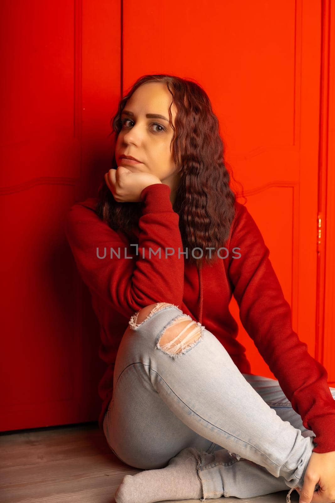 Young woman sitting on floor. Curly brunette poses near red wall