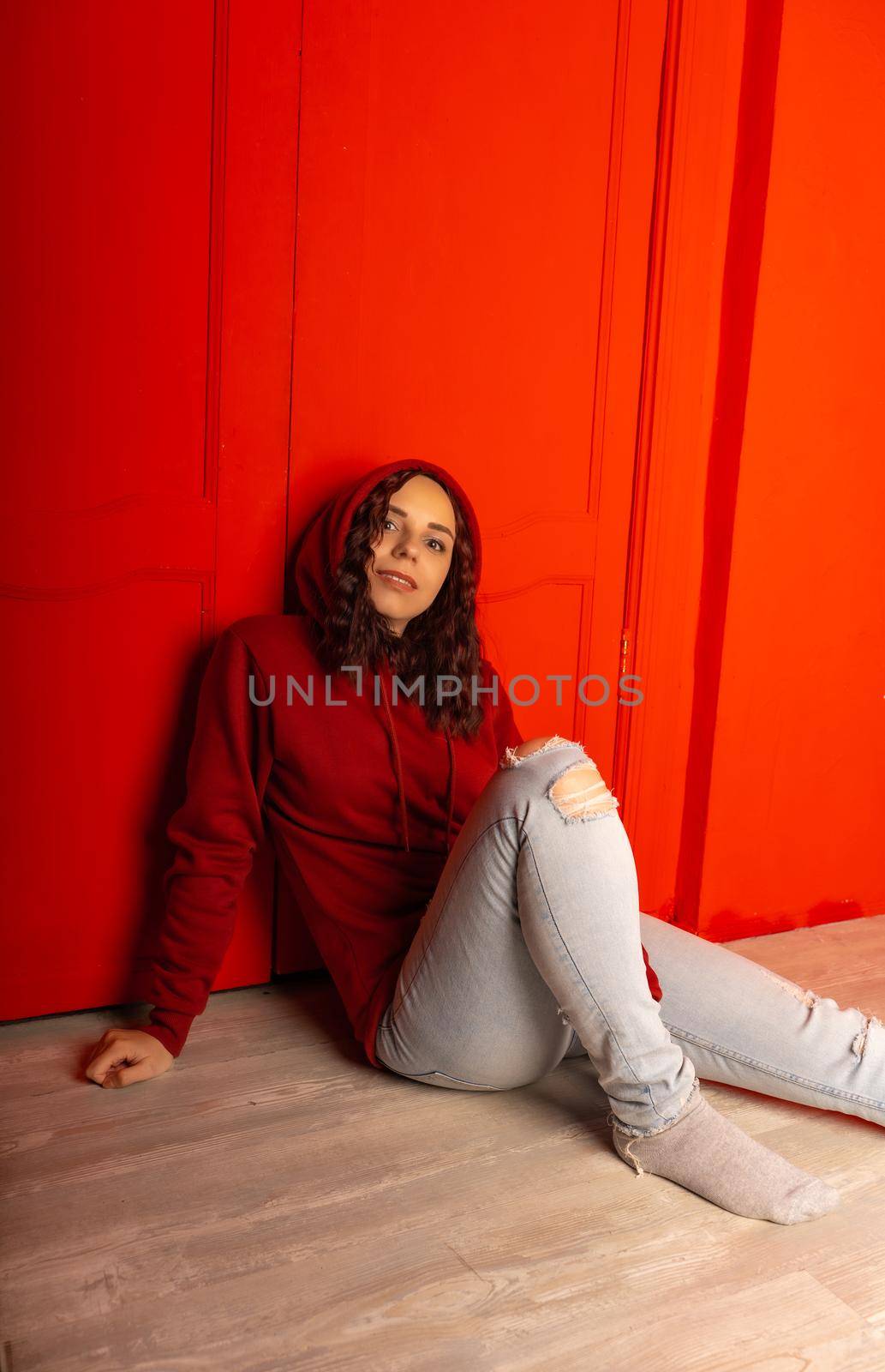 Young woman in hood sitting on floor. Curly brunette poses near red wall