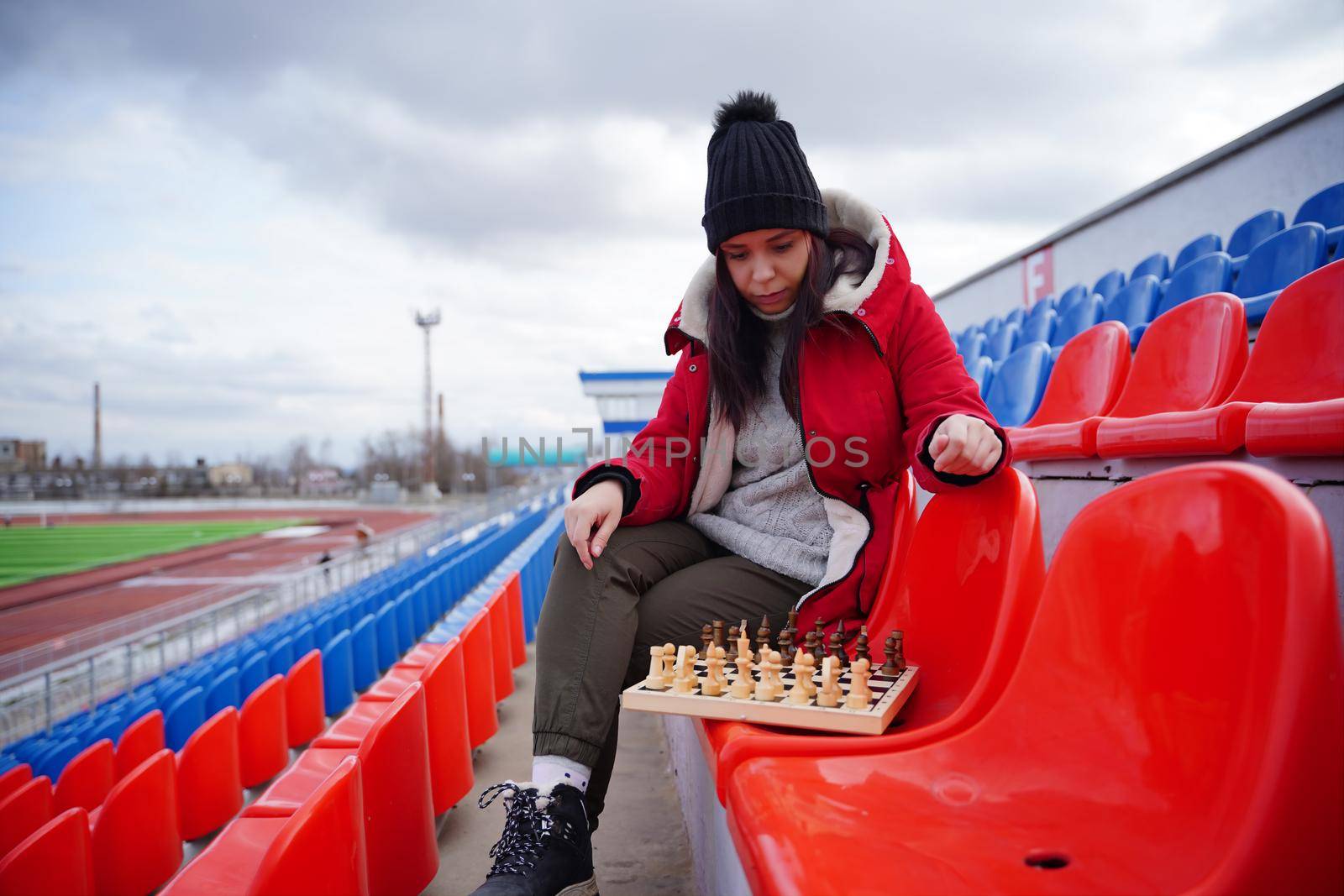 Young woman in winter clothes plays chess, sitting on stadium bleachers alone. Female in black cap with chess on sports stadium in cloudy weather. by epidemiks