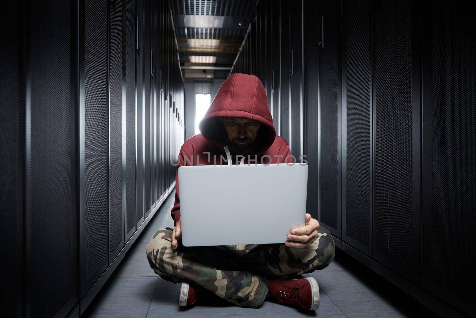 The hacker in a hood sits with the computer among servers, the burglar, face covered with a hood. Storage of data, cloudy services, system breaking, attack of hackers. High quality photo