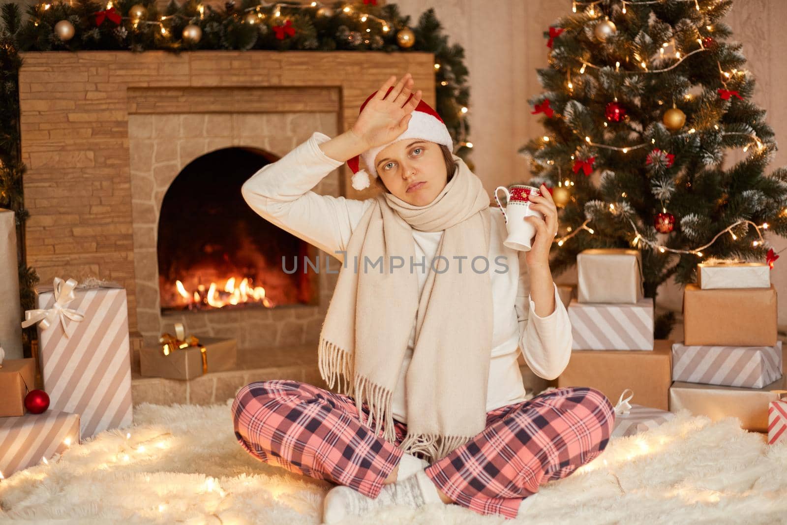 Young woman wrapped in warm scarf holds cup with hot tea, catches cold and flu during the festive winter holiday season, sitting on floor with crossed legs, touching her forehead.