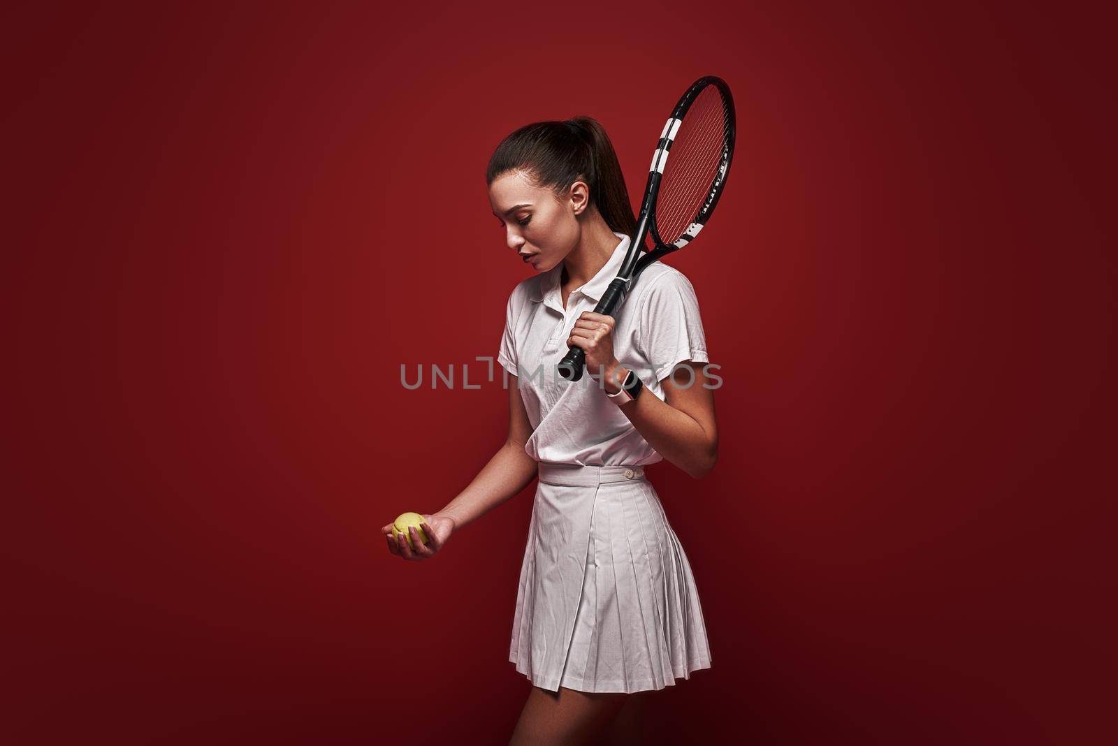Young tennis player in white polo shirt and skirt standing isolated over red background with a racket and a ball