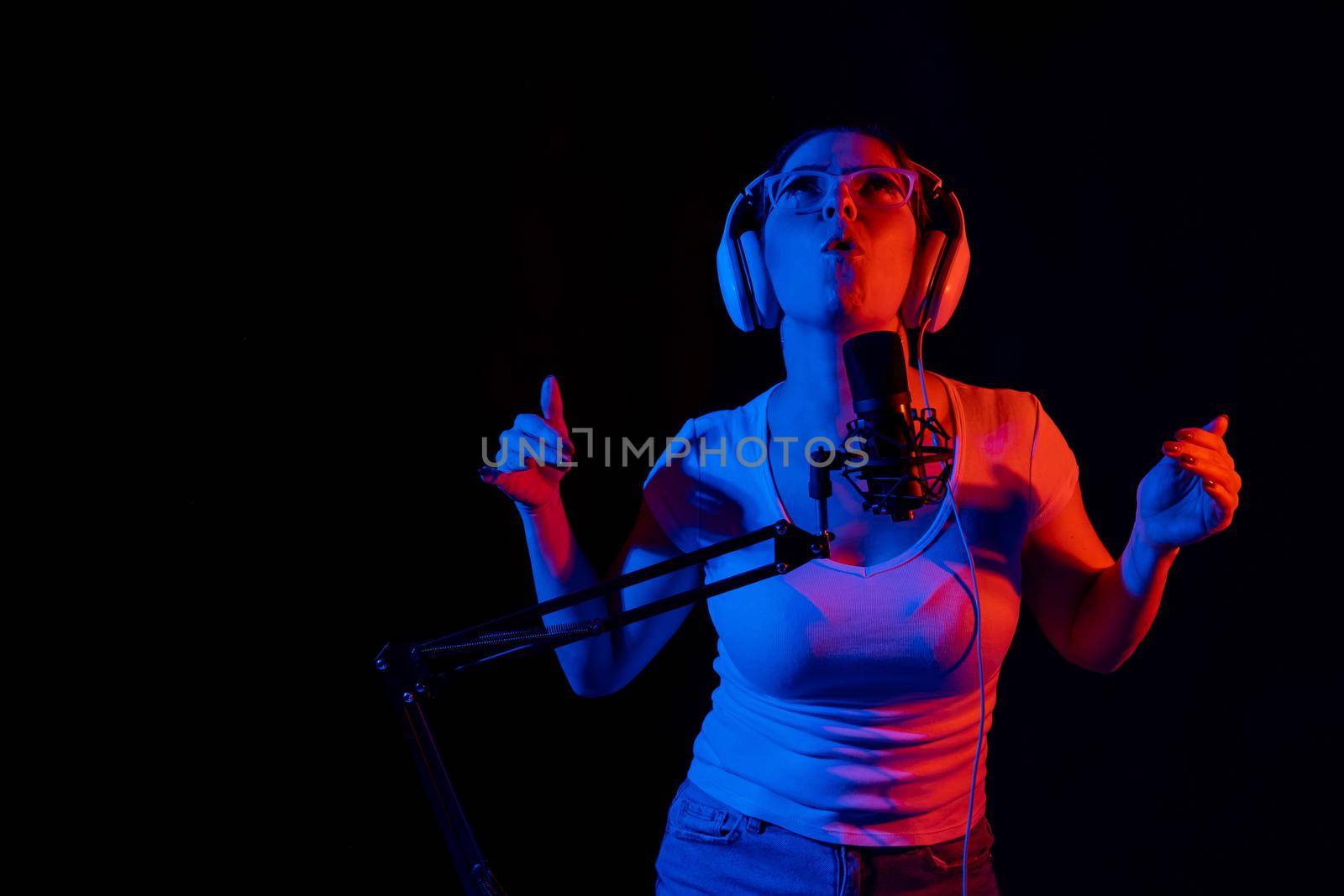 Caucasian woman in glasses and headphones sings into a microphone in neon light on a black background. An emotional girl is recording a song in a recording studio by mrwed54