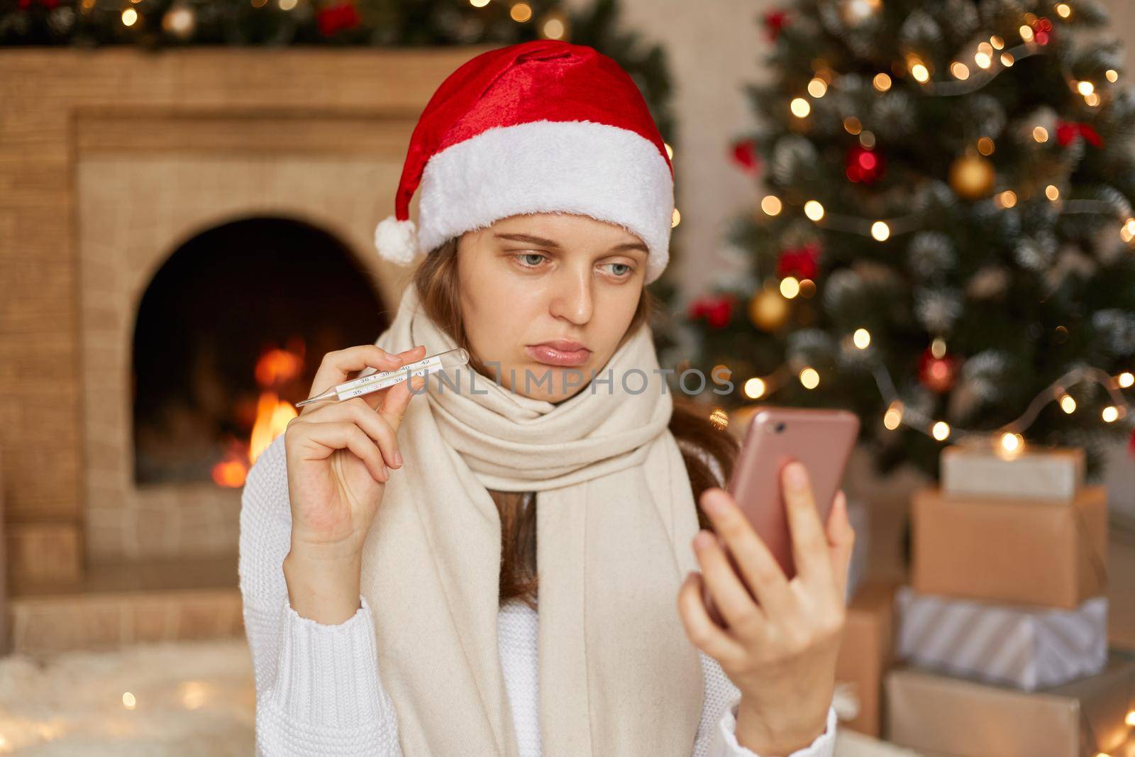 Unhappy and unhealthy woman posing in decorated living room phone in hands, talking via smart phone and showing thermometer to device camera, being disappointed to be ill during Christmas holidays.