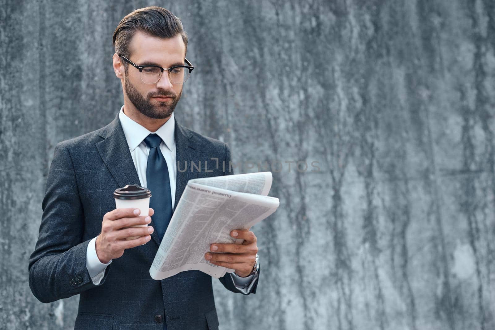 Young businessman in suit and glasses holding a paper cup and reading business newspaper in his hands