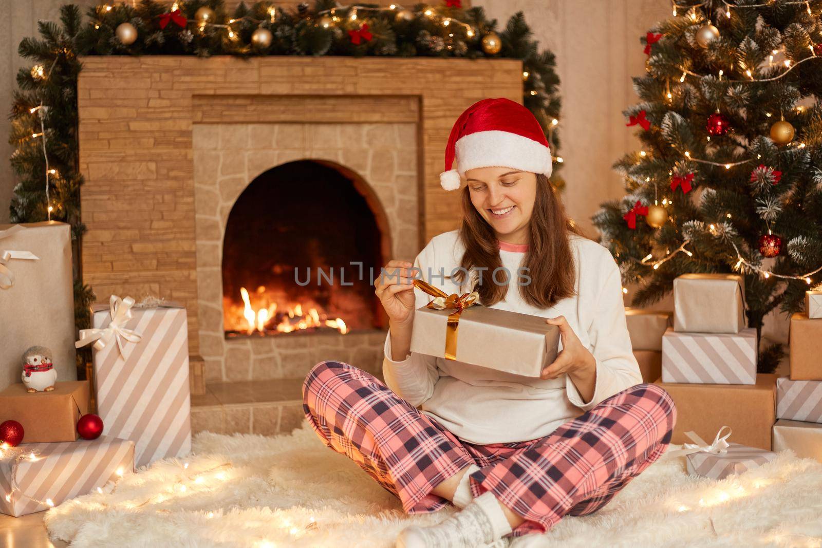 Caucasian woman opening present box while sitting on soft carpet in festive living room, lady wearing checkered pants, white shirt and santa claus hat, celebrating Christmas at home.