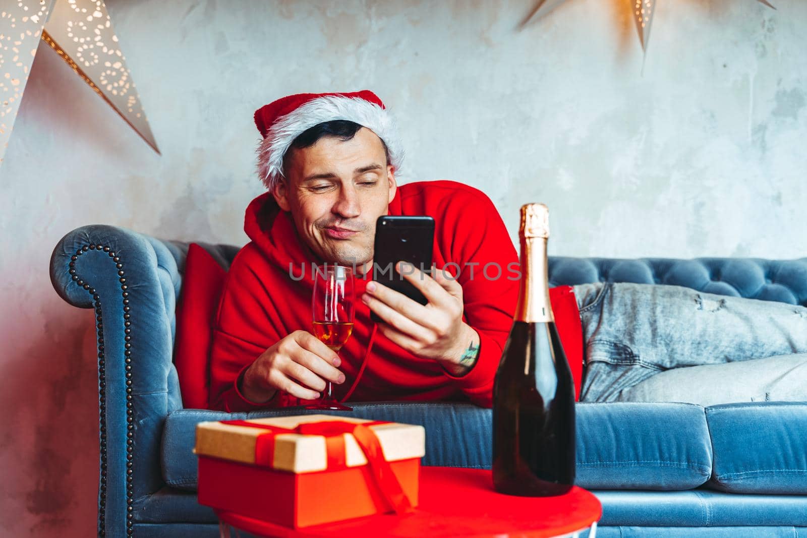 Young handsome man in Santa Claus hat with glass of champagne taking selfie on smartphone. Funny guy photographing himself on mobile phone and lying on couch. Concept of Christmas celebration at home