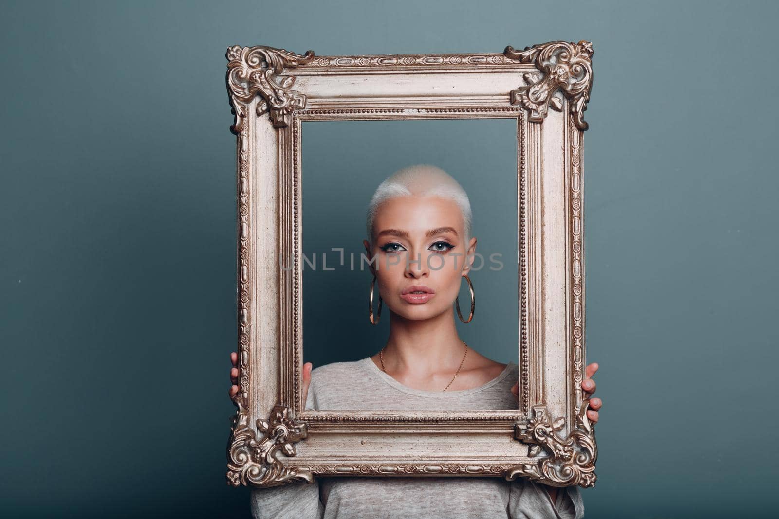 Millenial young woman with short blonde hair holds gilded picture frame in hands behind her face portrait