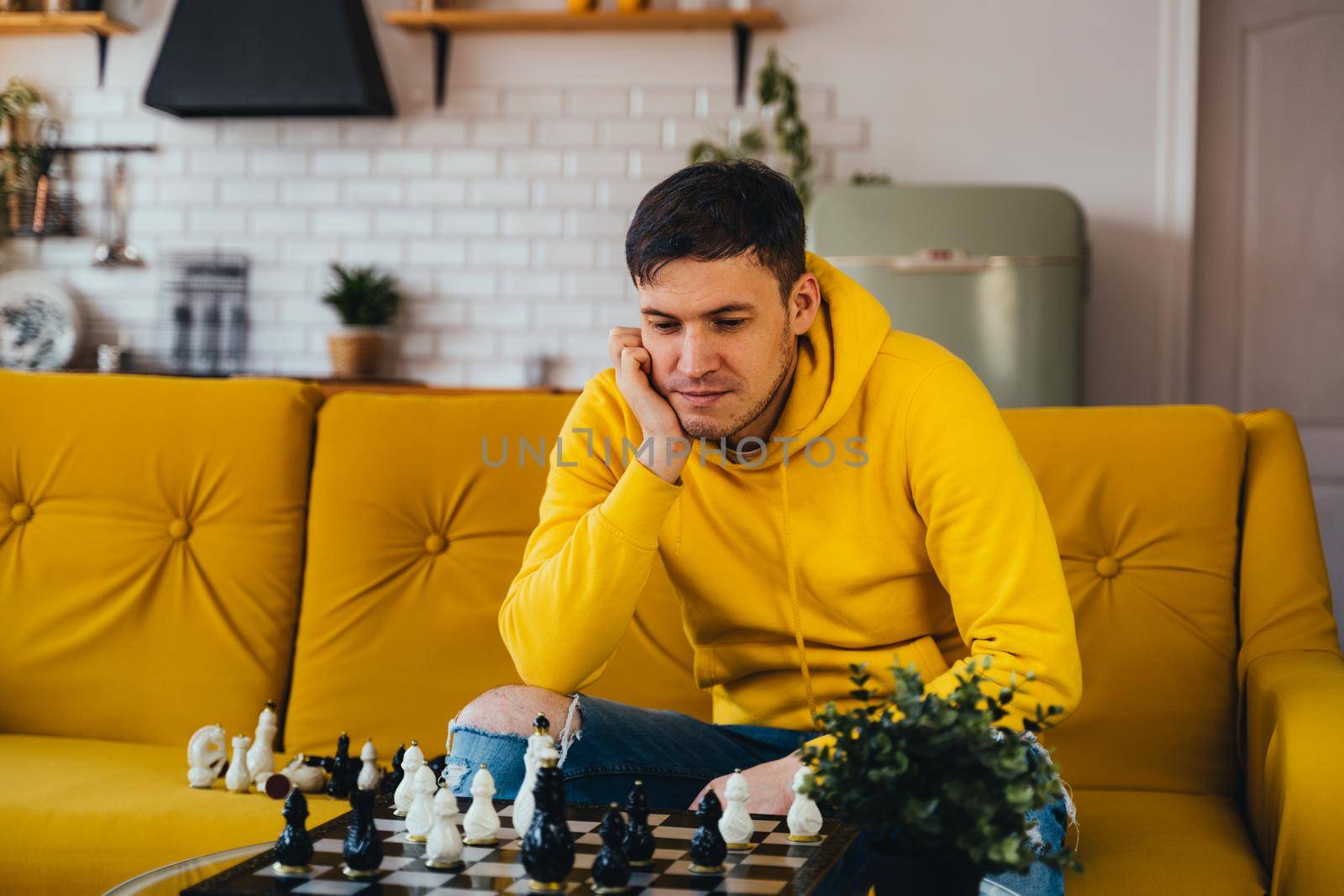 Young man sitting on yellow sofa and playing chess in room. Male playing in logical board game with himself. by epidemiks